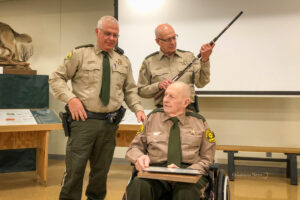 Mahaska County Sheriff Russ Van Renterghem (left), Dennis Dursky (center holding rifle), and Verlan Uitermarkt (seated) talk during a ceremony recognizing the years of service by Dursky and Uitermarkt on Sunday afternoon.