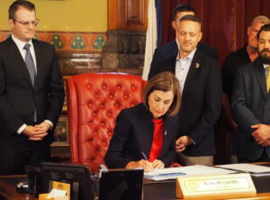 Gov. Kim Reynolds, joined by Lt. Gov. Adam Gregg, left, Sen. Dan Dawson, right, and other lawmakers and lobbyists, signed into law on May 1, 2024, a bill that will reduce Iowa’s individual income tax rate in 2025. (Photo by Robin Opsahl/Iowa Capital Dispatch)