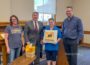 Mayor David Krutzfeldt, second from left, Oskaloosa Library Director Marion Gaughan, holding the lid, and City Manager Shawn Metcalf, right, during the America 250 Time Capsule sealing ceremony on July 21, 2026. The capsule contains 22 envelopes submitted by local residents and organizations and is scheduled to be opened July 4, 2076.